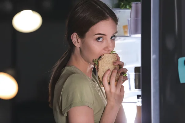 Wary young woman eating food near refrigerator at night — Stock Photo, Image