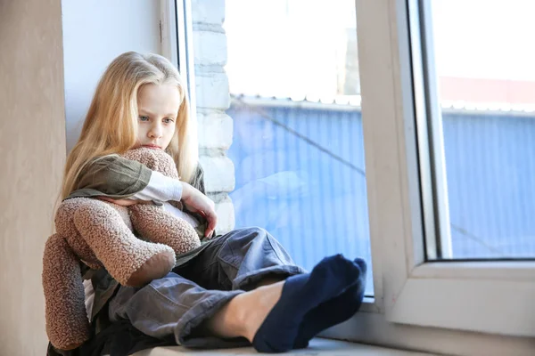 Бездомная маленькая девочка с плюшевым мишкой сидит на подоконнике — стоковое фото