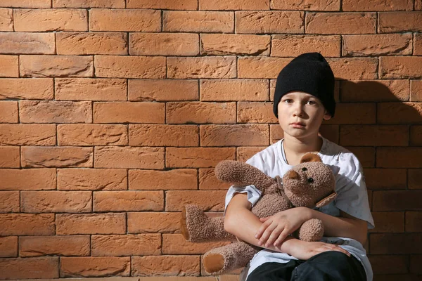 Niño sin hogar con oso de peluche sentado cerca de la pared de ladrillo — Foto de Stock