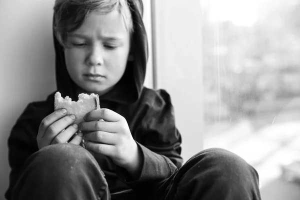 Niño sin hogar con pan sentado en el alféizar de la ventana dentro — Foto de Stock