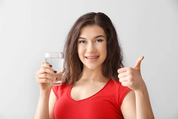 Beautiful young woman with glass of water showing thumb-up on light background — Stock Photo, Image