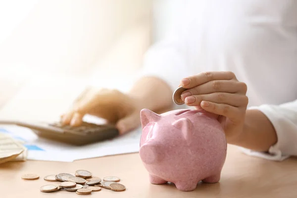 Young woman putting coin in piggy bank on table, closeup — Stock Photo, Image