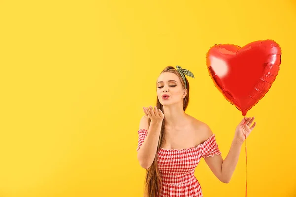 Mulher bonita com balão de ar em forma de coração soprando beijo no fundo de cor — Fotografia de Stock