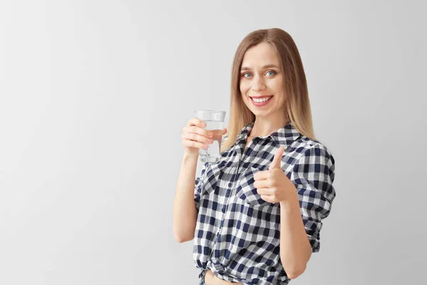 Mooie jonge vrouw met glas water dat duim-omhoog gebaar op lichte achtergrond toont — Stockfoto