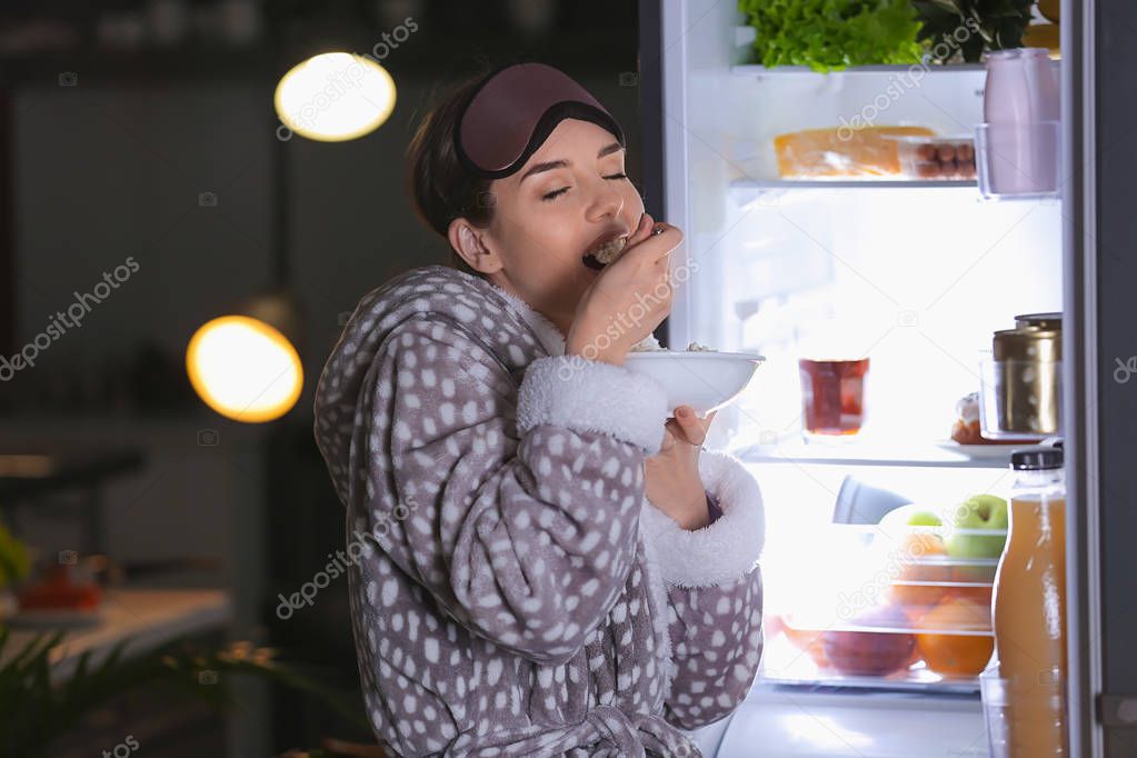 Beautiful young woman eating food near refrigerator at night