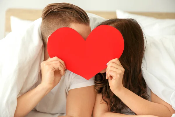 Feliz jovem casal com papel coração deitado na cama — Fotografia de Stock