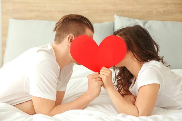 Glückliches junges Paar mit Papierherz im Bett liegend — Stockfoto