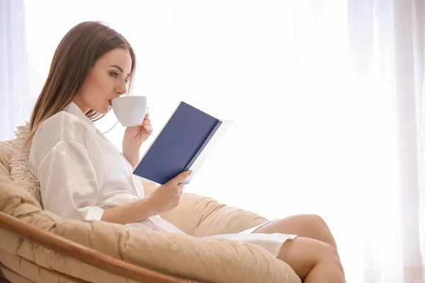 Piękna młoda kobieta pije kawę podczas czytania książki w domu — Zdjęcie stockowe