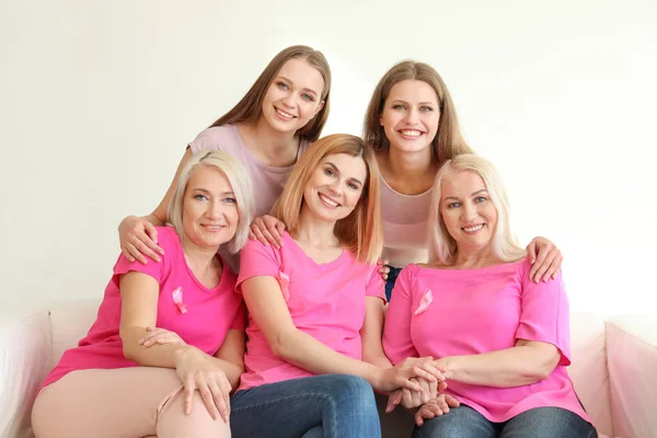 Hermosas mujeres de diferentes edades con cintas rosadas sentadas en el sofá en la habitación. Concepto de cáncer de mama — Foto de Stock