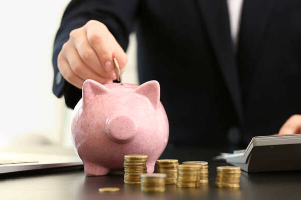 Woman putting coin into piggy bank and counting savings at table