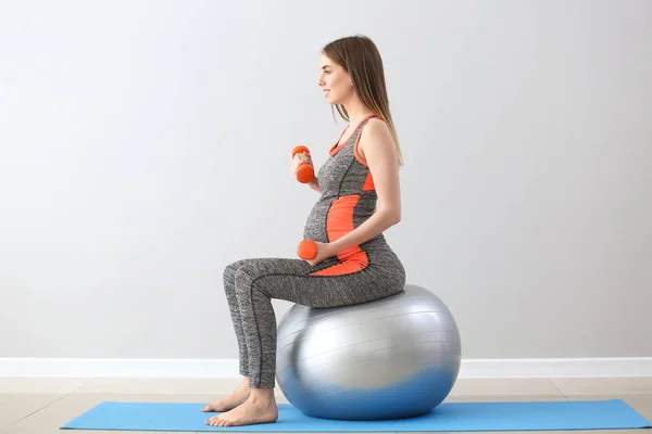 Mulher grávida bonita com halteres e treinamento fitball perto da parede cinza — Fotografia de Stock
