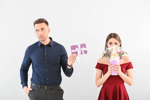 Displeased young man with gift box and happy woman with bouquet of beautiful flowers on light background — Stock Photo, Image