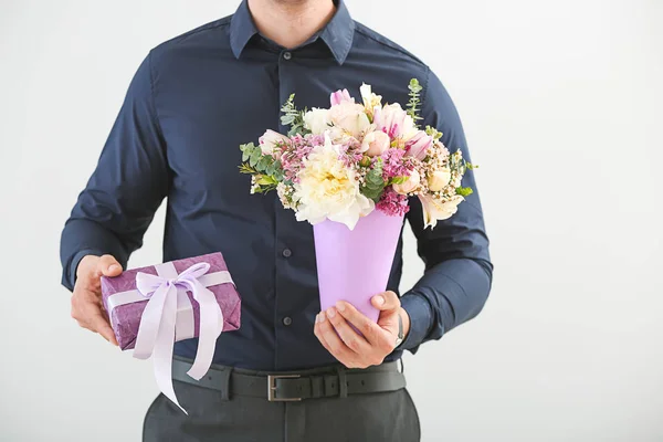 Handsome man with bouquet of beautiful flowers and gift box on light background
