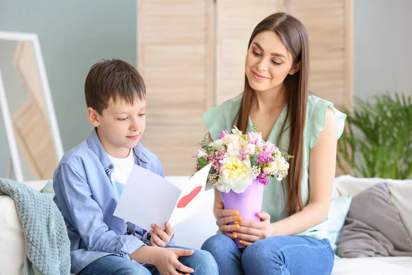 Little son greeting his mother with bouquet of flowers and greeting card at home — Stock Photo, Image