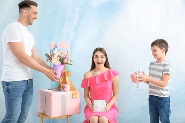 Surprised woman receiving gifts from husband and son on color background