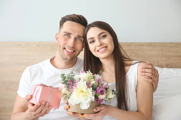 Gelukkig jong paar met gift box en boeket van bloemen zittend op bed thuis — Stockfoto