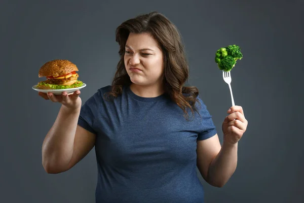 Indignant plus size vrouw met gezonde en ongezonde voedsel op grijze achtergrond. Concept van gewichtsverlies — Stockfoto