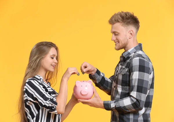 Casal feliz colocando moedas no banco porquinho no fundo de cor — Fotografia de Stock