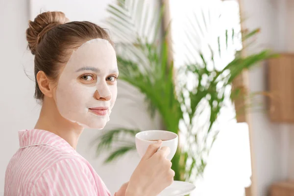 Jonge vrouw met blad gezichtsmasker drinken van thee thuis — Stockfoto