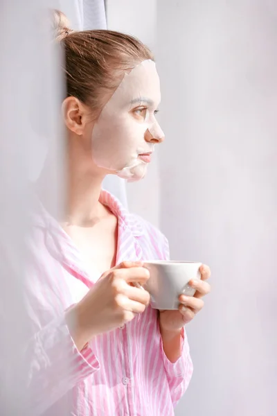 Mujer joven con sábana mascarilla facial bebiendo té en casa —  Fotos de Stock