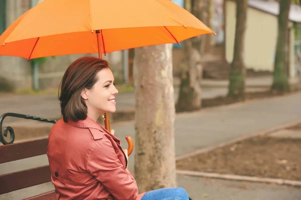 Hermosa mujer joven con paraguas sentado en el banco al aire libre — Foto de Stock