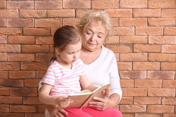 Linda niña con la abuela leyendo libro contra el fondo de ladrillo — Foto de Stock
