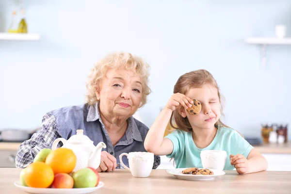 Cute little girl and grandmother drinking tea with cookies in kitchen — Stock Photo, Image