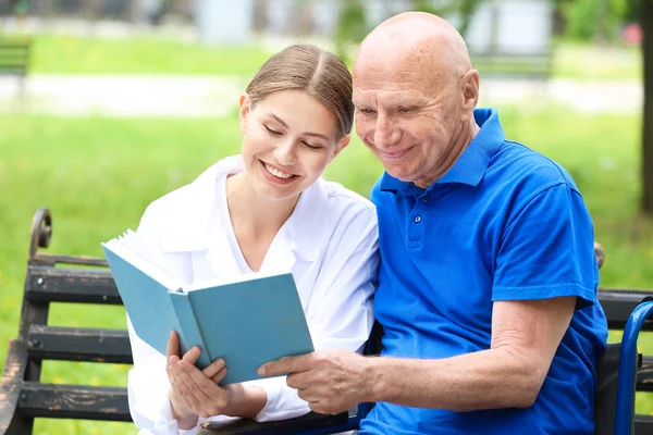 Uomo anziano con caregiver lettura libro nel parco — Foto Stock