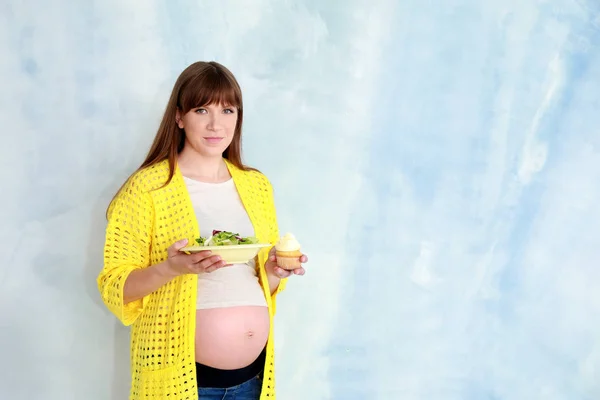 明るい背景にサラダとカップケーキを持つ美しい妊娠中の女性。健康食品と不健康食品の選択の概念 — ストック写真