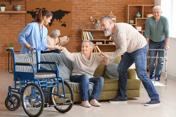 Caregiver and elderly man helping senior woman to sit in wheelchair — Stock Photo, Image