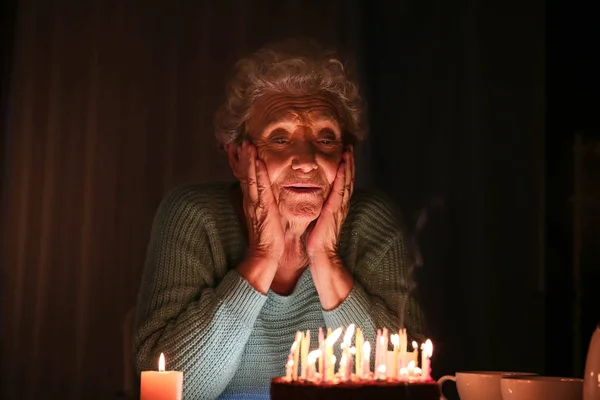 Lonely senior woman with birthday cake in dark room