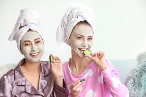 Hermosas mujeres jóvenes con máscaras faciales y rodajas de pepino en casa — Foto de Stock