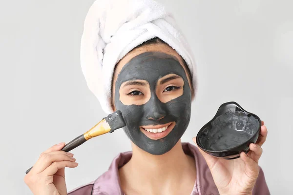 Beautiful Asian woman applying facial mask against light background — Stock Photo, Image