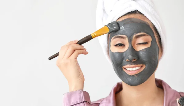 Mooie Aziatische vrouw gezichtsmasker toepassen tegen lichte achtergrond — Stockfoto