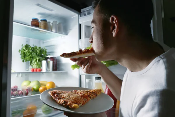 Молодий чоловік їсть піцу біля холодильника вночі — стокове фото