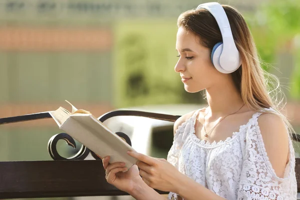 Beautiful young woman listening to music while reading book in park — Stock Photo, Image