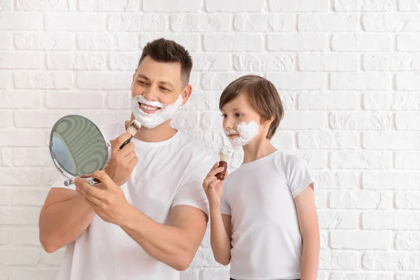 Father and his little son with shaving foam on faces against white brick wall — Stock Photo, Image