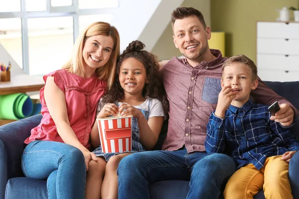 Casal feliz com crianças adotivas comendo pipocas enquanto assiste TV em casa — Fotografia de Stock