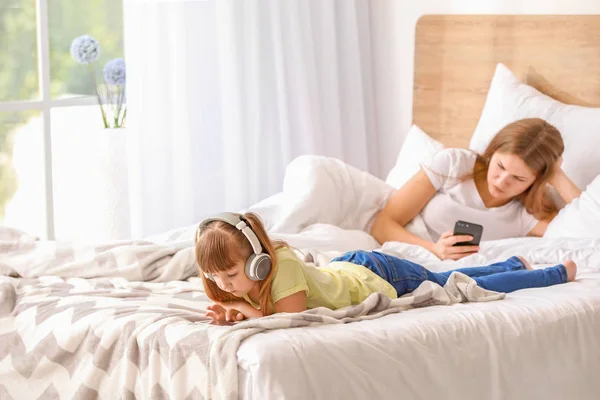 Little girl and mother addicted to modern technologies with devices on bed at home — Stock Photo, Image