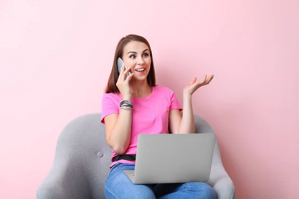 Beautiful young woman with laptop talking by phone while sitting in armchair on color background — Stock Photo, Image