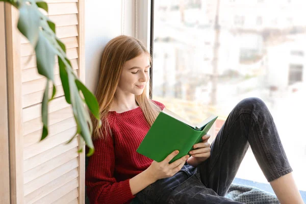 Linda adolescente leyendo libro en alféizar ventana en casa — Foto de Stock