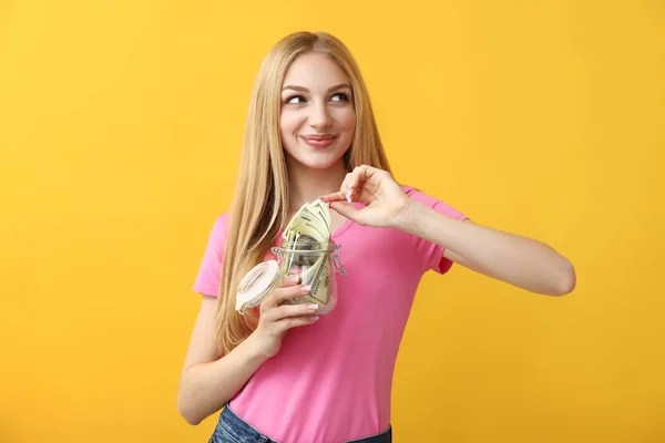 Feliz joven mujer sosteniendo frasco de vidrio con dinero en el fondo de color — Foto de Stock