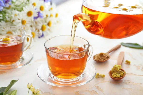Pouring of tasty chamomile drink from teapot into cup on light table — Stock Photo, Image