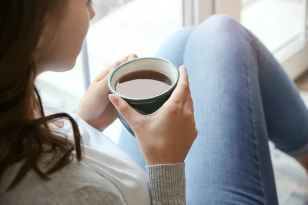 Mooie jonge vrouw drinken warme koffie in de buurt van venster thuis — Stockfoto