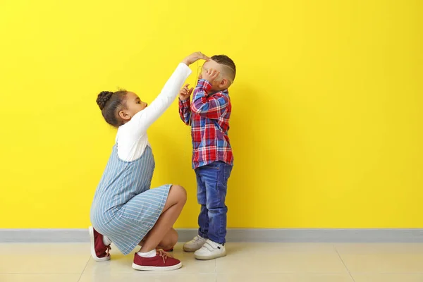 Cute little African-American girl measuring height of her brother near color wall — Stock Photo, Image