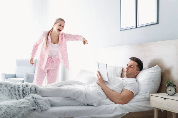 Злая молодая женщина кричит на своего мужа, который читает книгу в постели — стоковое фото