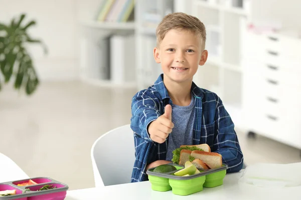 Little schoolboy with tasty lunch in box showing thumb-up gesture in classroom — Stock Photo, Image