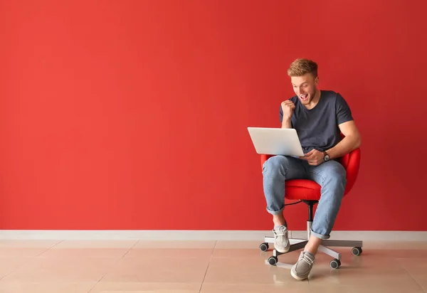 Happy young man with laptop sitting on chair near color wall — Stock Photo, Image