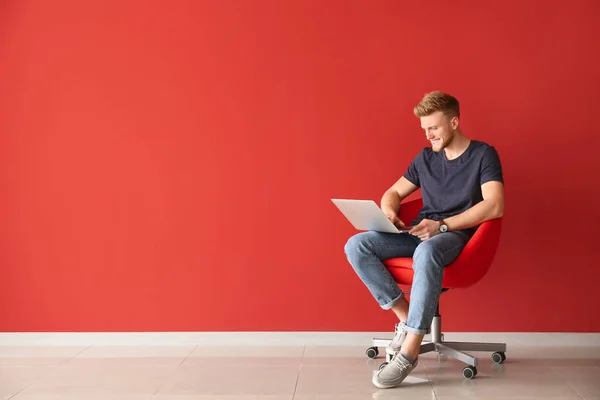 Handsome young man with laptop sitting on chair near color wall — Stock Photo, Image