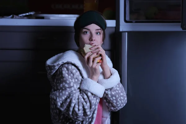 Wary young woman eating unhealthy food near fridge in kitchen at night — Stock Photo, Image